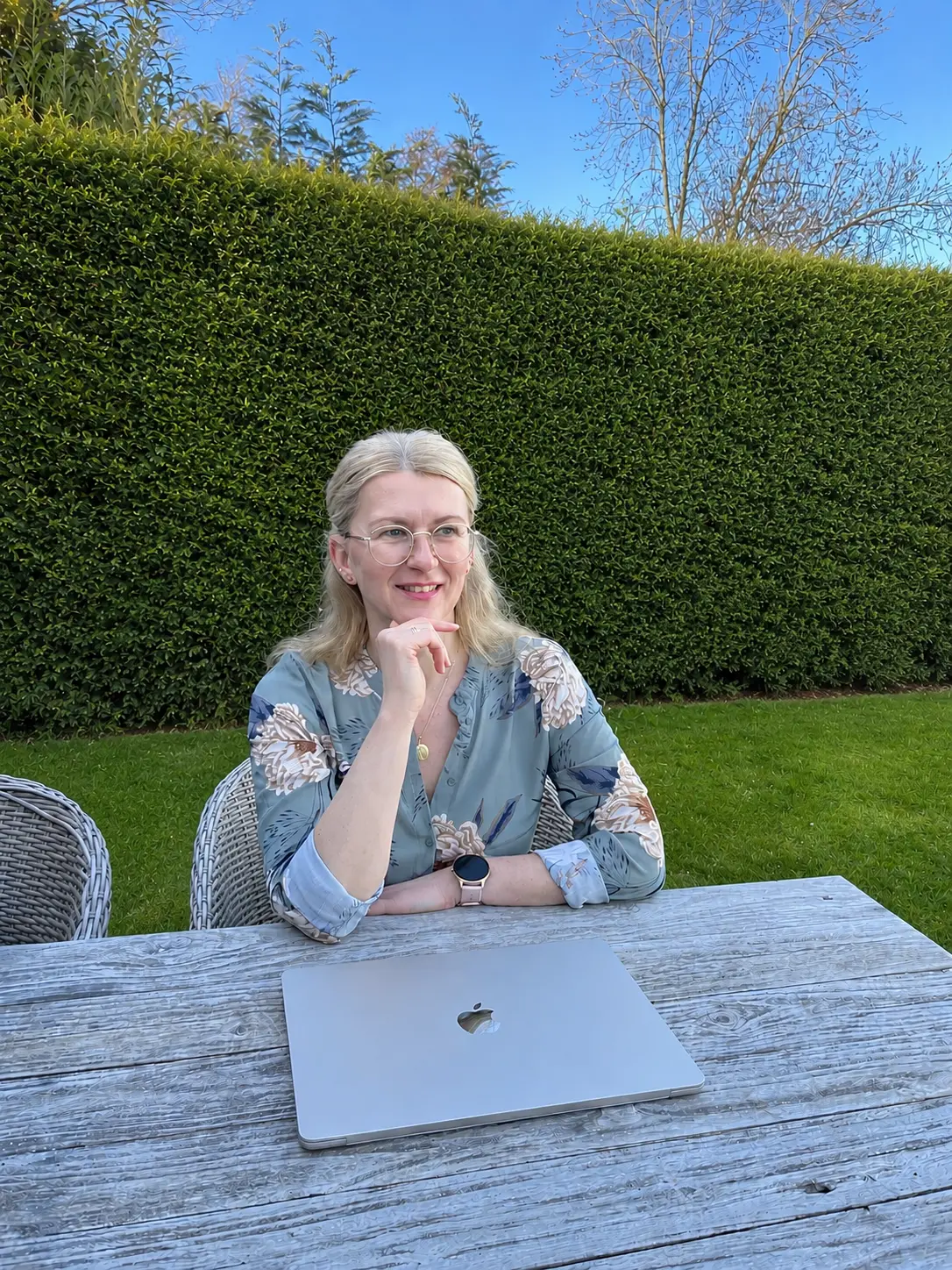 Isabella Örtülü, Gründerin der isi-netzwerkagentur, lächelnd und nachdenklich an einem Holztisch im Garten sitzend. Vor ihr liegt ein MacBook; im Hintergrund ist eine hohe Hecke zu sehen. Sie trägt eine florale Bluse und eine Smartwatch.