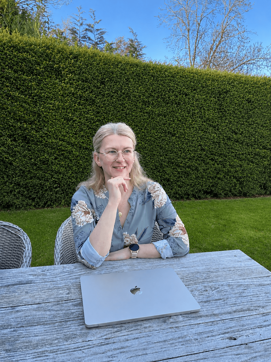 Isabella Örtülü, Gründerin der isi-Netzwerkagentur, lächelnd und nachdenklich an einem Holztisch im Garten sitzend. Vor ihr liegt ein MacBook; im Hintergrund ist eine hohe Hecke zu sehen. Sie trägt eine florale Bluse, Brille und eine Smartwatch.