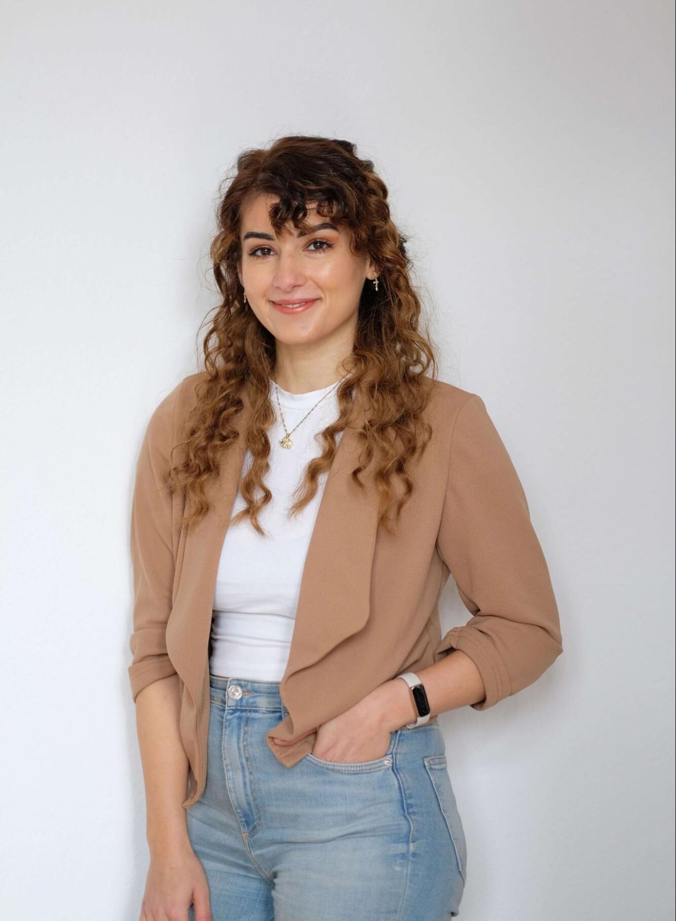 Porträt von Aylin Nil Örtülü, Social Media Managerin und Freelance Content Strategin, lächelnd mit lockigem Haar vor einem hellen Hintergrund. Sie trägt einen braunen Blazer, ein weißes Shirt und Jeans. Netzwerkpartner der isi-Netzwerkagentur.