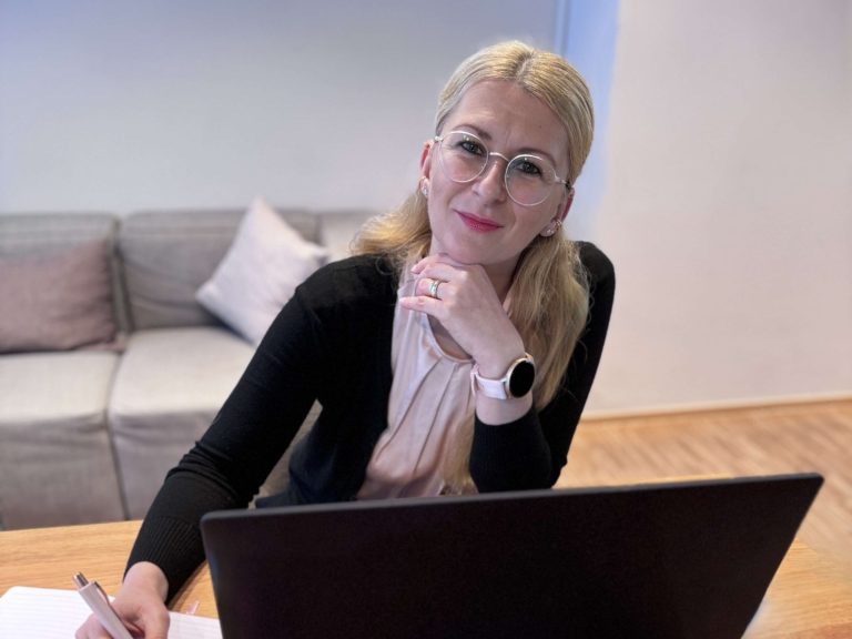 Frau mit Brille sitzt an einem Tisch und arbeitet am Laptop, Lächeln im Gesicht.