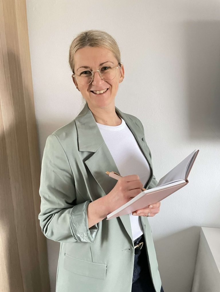 Eine lächelnde Frau mit Brille hält ein Notizbuch und einen Stift in der Hand.
