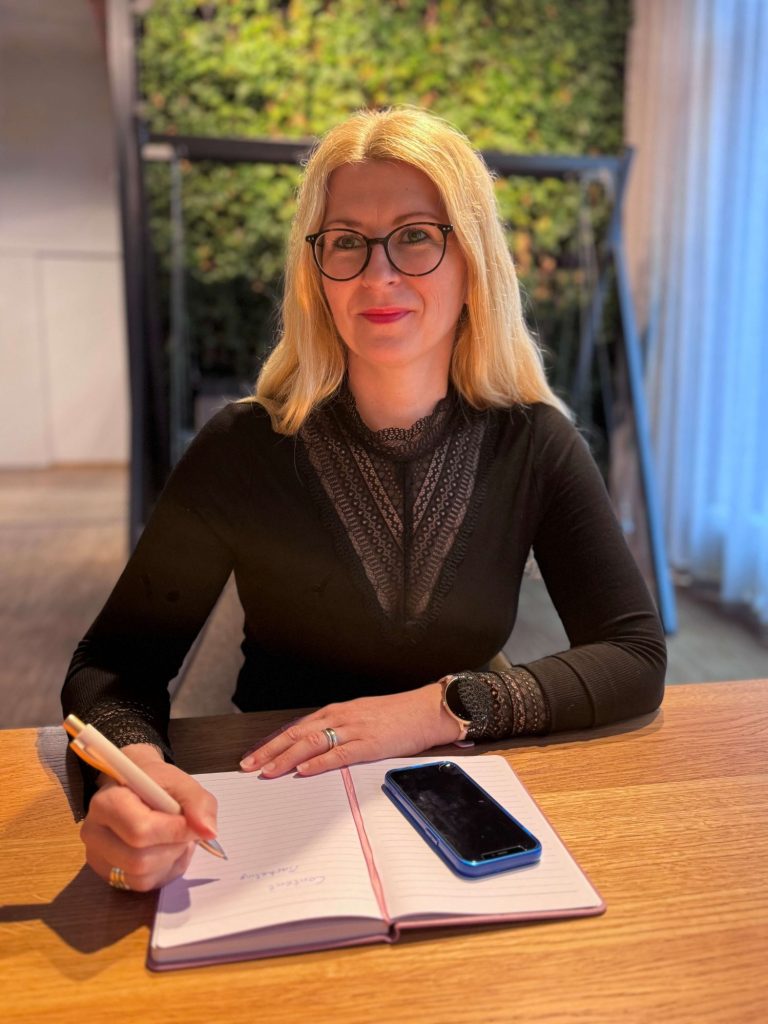 Frau mit blonden Haaren und Brille, die in ein Notizbuch schreibt, Smartphone daneben.