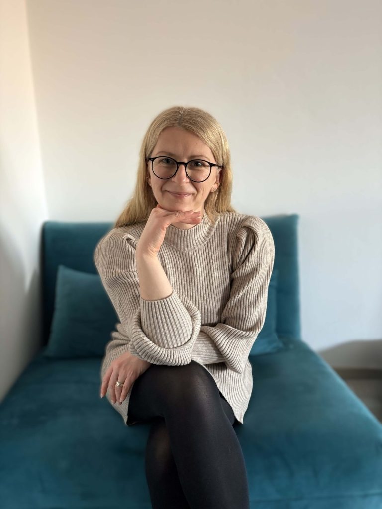 Frau mit langen blonden Haaren sitzt auf einem blauen Sofa und lächelt freundlich.